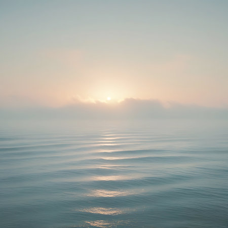 The calm surface of the ocean displays delicate, repeating wave patterns. Soft sunlight from a hazy sunrise reflects brightly on the water, creating a shimmering path towards the horizon. The sky is a blend of pale blues and soft pinks.の素材
