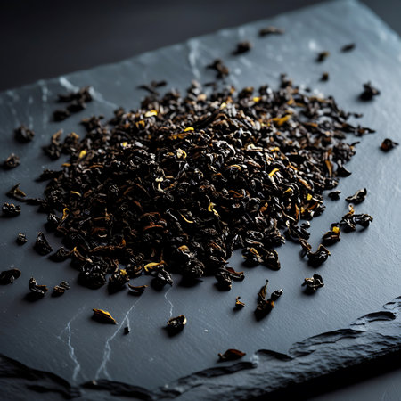 A close-up, top-down view of a pile of dark, loose leaf tea scattered across a textured, dark grey slate surface. The tea leaves are a deep black with hints of yellow and brown, appearing dry and brittle. The slate has subtle white veining, creating a sophisticated and minimalist presentation.の素材