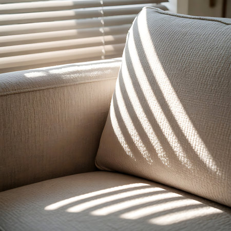 This image focuses on the corner of a light-colored sofa, specifically a textured pillow and the armrest. Diagonal stripes of sunlight, cast by window blinds, create a striking visual pattern across the fabric. The detail of the woven texture is emphasized by the interplay of light and shadow, suggesting a warm and inviting interior space.の素材