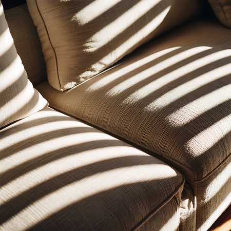 A close-up view of a textured fabric sofa with distinct stripes of sunlight cast across its cushions and armrest. The light, originating from a window with blinds, creates a geometric pattern of light and shadow, highlighting the woven texture of the upholstery. The overall impression is one of cozy domesticity and the play of natural light.の素材