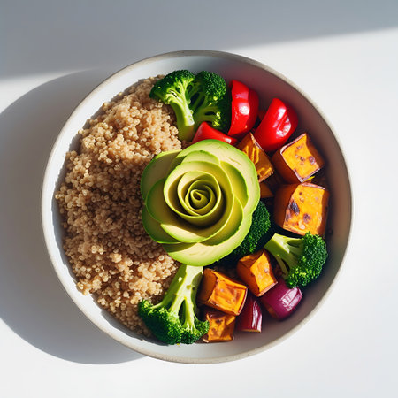 This image is a top-down view of a healthy quinoa bowl. A meticulously carved avocado forms a rose shape in the center. The quinoa is encircled by roasted sweet potato cubes, broccoli florets, red bell pepper slices, and red onion pieces. The bowl rests on a plain white surface, illuminated by bright light that creates subtle shadows, emphasizing the textures and colors of the fresh ingredients.の素材