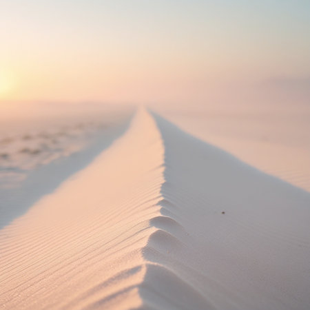The image captures a tranquil scene of sand dunes bathed in the soft, warm hues of a setting sun. The sand appears smooth and undulating, with gentle ripples created by the wind. The sky transitions from a light orange near the horizon to a soft pink and finally to a pale blue as it stretches upwards. The overall mood is peaceful and serene, evoking a sense of calm and solitude.の素材