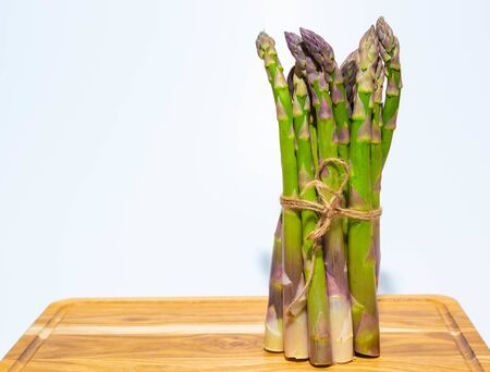 Asparagus standing upright on a wooden board on a white background. A bunch of asparagus shoots tied with a rope on a kitchen cutting board on a white background.の写真素材