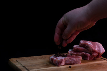 Piece of pork brisket on a kitchen board on a dark background. The cook's hand sprinkles allspice over the raw meat.の写真素材
