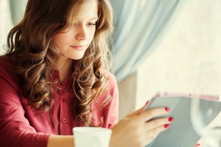 Young smiling woman is drinking coffee in a cafe and holding a tablet computer in handsの写真素材
