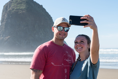 A young couple of tourists make selfies in front of the Haystack Rock in Cannon Beach, Oregon, USA.のeditorial素材