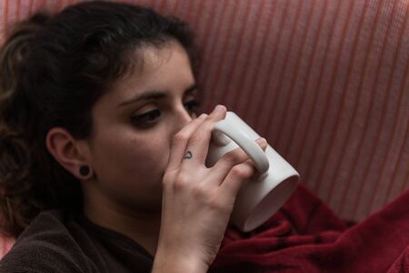 Dark-haired, thoughtful young woman takes a cup sitting on the couch.