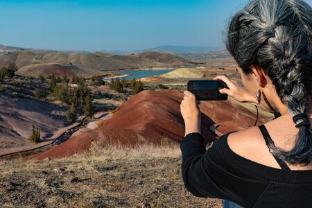 A young woman with dark hair and gray hair makes a photograph with her smartphone from the Painted Hill Reservoir, Oregon, USAの写真素材