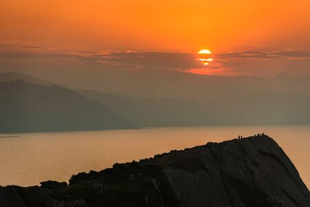 Tiny silhouettes of people watching the sunset from the top of a cliff in Zumaiaの写真素材