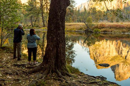 A couple takes pictures of the reflection of the landscape in the water of the Merced River in Yosemite National Parkのeditorial素材