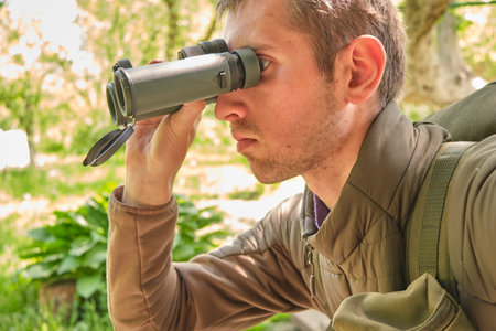 young soldier in olive military tactical gear looks into green tactical binoculars in the summer afternoon in the heat of the sunの写真素材