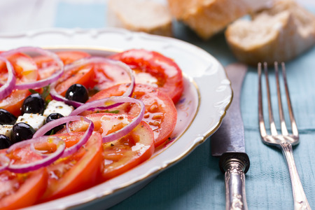 Tomato, Feta, Onions and Arugula Salad in a white bowl on white wooden background with antique fork and knifeの写真素材