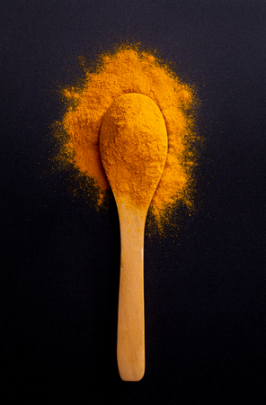 Heap of curcuma turmeric spice on wooden spoon on dark black background, top view