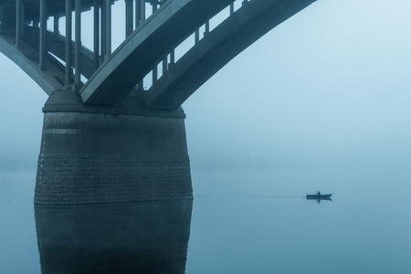 fisherman on a small boat sails next to the pillars of the bridge in the fog.の写真素材