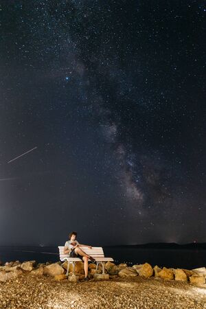 A young male traveler in shorts and a T-shirt resting on a bench over a beautiful clear sky with a milky way.の写真素材