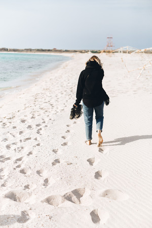 A young girl walks along an empty beach alone. Shoes in hand, footprints in the sandの写真素材