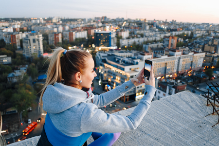 Young girl in sports uniform takes photos on the phone on the roof of a building over the city.の写真素材