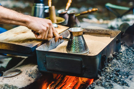 Man is cooking Turkish coffee in the box with sand under live coals near a riverの写真素材
