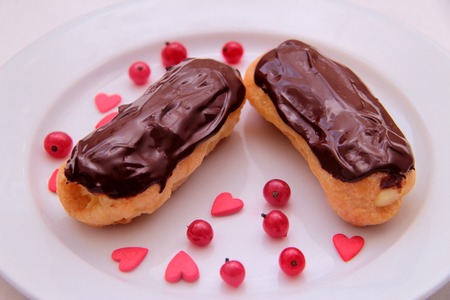 Chocolate eclairs on plate on white background sprinkled with red currant berries and hearts.
