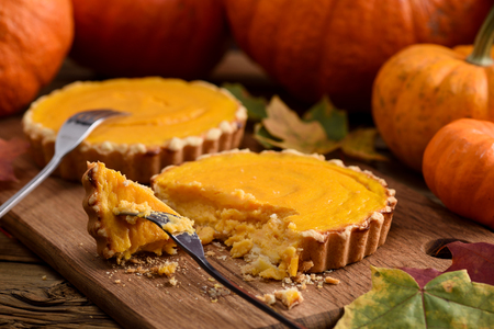Delicious small pumpkin pies being cut and eaten with forks on wooden board decorated with orange pumpkins closeupの写真素材