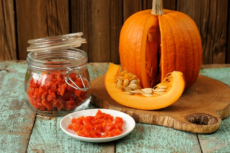 Homemade candied pumpkin pieces in glass jar and white plate with cut pumpkin with seeds on old wooden table horizontalの写真素材