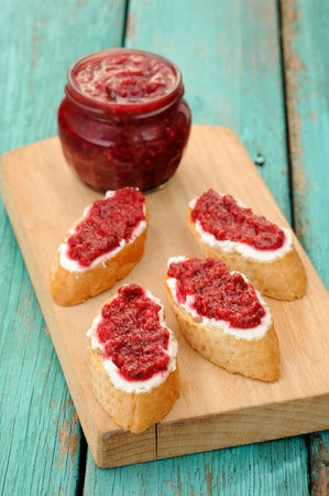 Homemade sweet raspberry jam in glass jar with four toasts with cream cheese and berry jam on wooden...