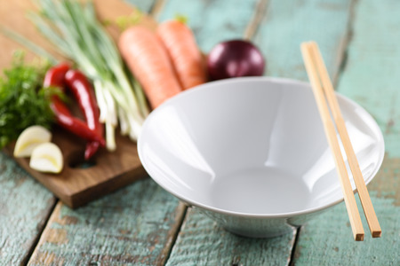 Empty white bowl and raw vegetables, ingredients for Asian udon noodles with chop sticks on wooden blue background closeupの写真素材