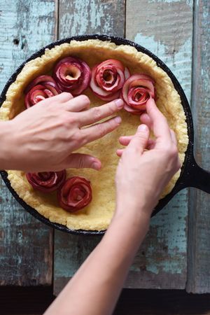 Homemade fruit pie. Woman hands making apple roses for pie in cast iron pan on shabby blue background overhead viewの写真素材