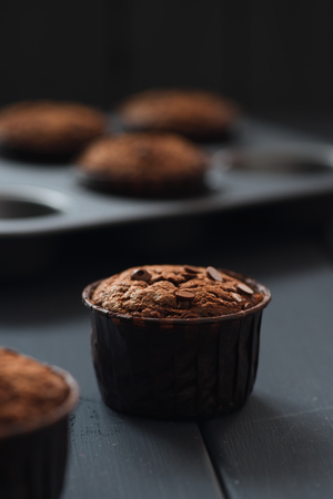 Minimalist style hygge dessert. Chocolate muffins with chocolate drops in brown paper and in baking tray on dark background verticalの写真素材