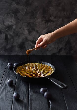 Slender woman hand with honey dipper decorating homemade flower shaped plum pie in cast iron pan on black background copy spaceの写真素材