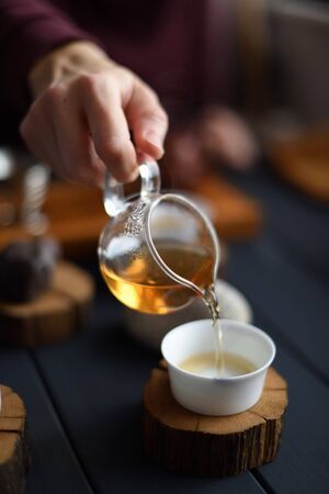 Healthy beverage. Slender woman hand pouring Chinese green tea from glass pitcher into porcelain cup on wood slab on dark background verticalの写真素材
