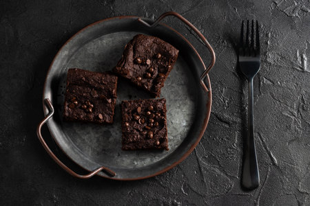 Dark chocolate brownies with choco drops on metal tray on dark plaster surfaceの写真素材