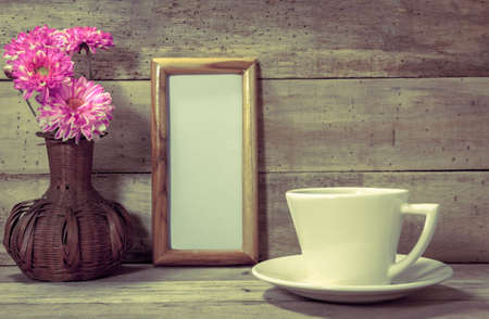 Selective focus cup of coffee,white paper space in picture frame,beautiful pink flower in jar on wood background by vintage tone.の写真素材
