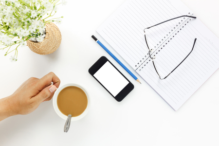 Top view business office desk.Hand Businessman touching cup of coffee and mobile phone,eyeglasses,coffee,notebook,pencil,beautiful white flower on white office desk with copy space.の写真素材