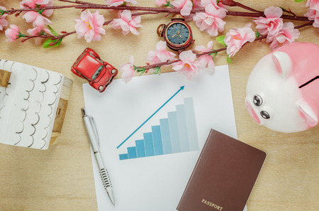 Top view business office desk concept. Saving money to travel with passport.charted summary with Wood house  also car and clock on wooden shelf.The beautiful pink flower with pen on wood background with copy space.の写真素材