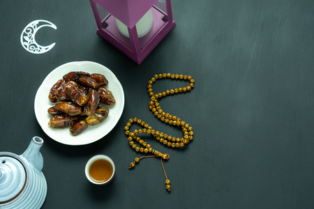 Table top view aerial image of decoration Ramadan Kareem holiday background.Flat lay date with rosary & lighting and cup of tea.Essential object on modern rustic black wooden at office desk.copy spaceの写真素材