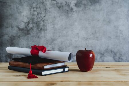 Close up of Graduation of education season background concept. Accessory cap with certificate paper & book and red apple on modern rustic brown wooden and grey cement.graduated of university.の写真素材