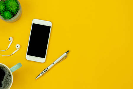 Table top view aerial image of accessories office desk background concept.Flat lay of variety object the white white mobile phone & tree pot and with earphone on modern rustic yellow paper and space.の写真素材