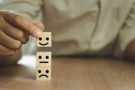 Close up Hand holding of Happy icon on cube blocks about service rating feedback of survey from customer. Business annual satisfaction survey concept. Many sad or happily on wooden table.copy spaceの写真素材