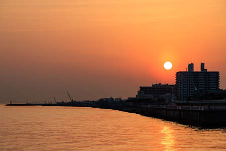 Sun sets through orange haze over silhouetted buildings on coastの写真素材