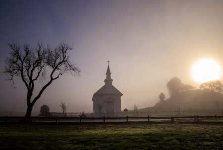 Bright sun and dark fog over small church and tree in rural countryside at dawnの写真素材
