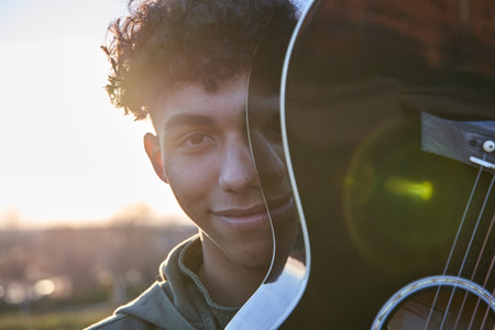 Latin teenager covering his face with guitar. Smiling young man with curly brown hair at sunset.の写真素材