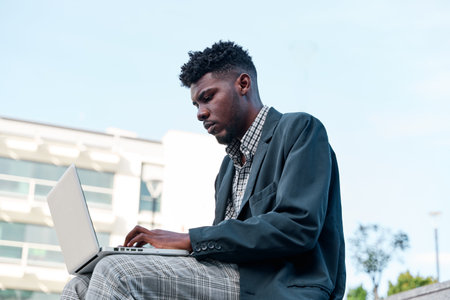 Side view of an African American man working with his laptop sitting on a staircase in the city, wearing casual clothes.の写真素材