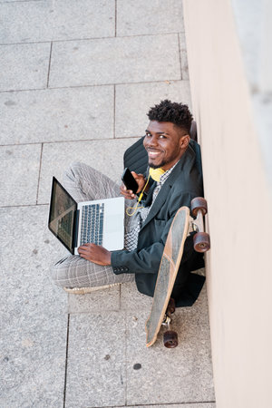 Young businessman sitting outdoors at a wall working on laptop looking at camera with smartphoneの写真素材