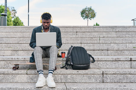 front view of an african american man sitting on a staircase working on his laptop. hipster man listening to music with yellow headphones.の写真素材