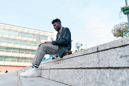 Side view of an African American man working with his laptop sitting on a staircase in the city, wearing casual clothes.の写真素材