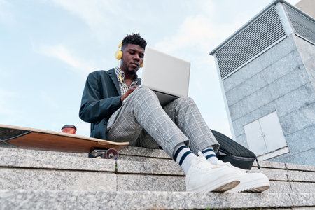 View from below of a young African-American adult man on a skateboard while working on a laptop computerの写真素材