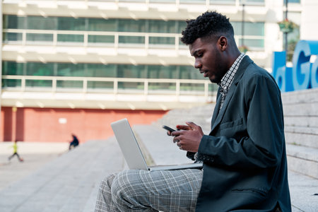young African-American man sitting on a city staircase with his laptop while using his mobile phone.の写真素材