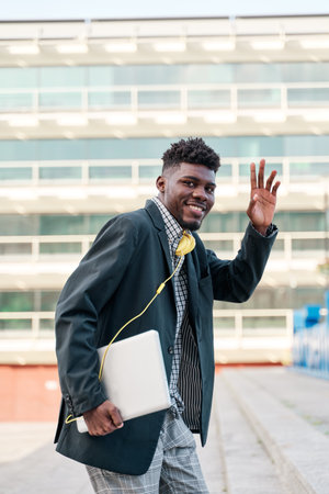 Happy african american man dressed in casual denim jacket waving hand to greet friend while walking with modern laptop computer in hands outdoors on street.Positive hipster guy with netbook gesturing helloの写真素材