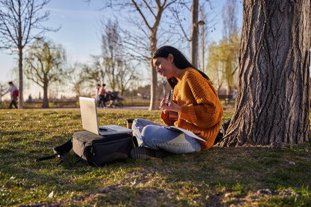 young latina with long hair using a laptop while playing the ukulele under a tree in a park.の写真素材